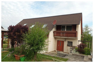 Dvojpodlažný dom s KATEPAL Oy Asfaltový strešný šindeľ Katepal 3T - hneďý strecha, červené vchodové dvere, balkón, satelitná anténa a strešné okná. Na dvore sú stromy, kríky a zelená záhradná hadica pod čiastočne zamračenou oblohou.