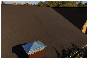 Detailný záber na šikmú strechu s Asfaltovým strešným šindeľom Katepal 3T - hneďý od spoločnosti KATEPAL Oy a komín s kovovou krytinou v slnečnom svetle, v pozadí vidno stromy a modrú oblohu.