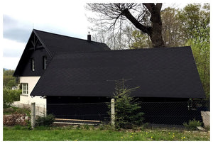 Čierny dom so strmými strechami pokrytými Asfaltovým strešným šindeľom Katepal Foxy - čierny (KATEPAL Oy) je čiastočne ukrytý za zodpovedajúcou garážou, obklopený stromami, kríkmi a drôteným plotom pozdĺž trávnatého priečelia.