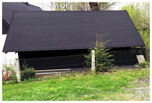 Veľký otvorený prístrešok s reťazovým plotom, malými vždyzelenými stromami, lavičkou a zelenou trávou pred ním. Čierna strecha je pokrytá Asfaltovým strešným šindľom Katepal Foxy - čierny od spoločnosti KATEPAL Oy. V pozadí je vidieť stromy a kríky.