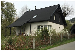 Domček krémovej farby s čiernym Asfaltový strešný šindeľ Katepal Foxy - čierny od spoločnosti KATEPAL Oy stojí pri ceste, obklopený zeleňou a stromami, s malou terasou a vonkajším posedením pri vchode.