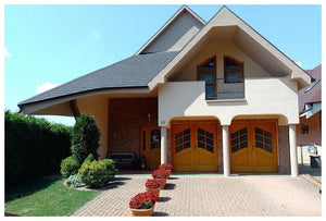 Moderný dom so svetlými stenami, dvoma drevenými garážovými bránami a oblúkovou strechou pokrytou KATEPAL Oy Asfaltový strešný šindeľ Katepal Mansion - hneďý. Červené kvety v kvetináčoch lemujú príjazdovú cestu; naľavo sa rozprestiera malá záhrada so živým plotom a kríkmi.