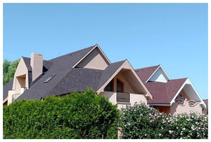 Rad moderných domov so šikmé strechy pokryté hnedý asfaltový šindeľ Katepal Mansion od KATEPAL Oy, béžové steny, a veľké trojuholníkové okná, čiastočne skryté zelenými kríkmi a kvitnúce kríky pod jasnou modrou oblohou.