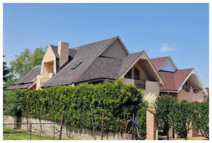 Dva moderné domy so šikmými strechami pokrytými KATEPAL Oy asfaltový strešný šindeľ Katepal Mansion - hneďý sú čiastočne ukryté za vysokým zeleným živým plotom. Pozdĺž priečelia sa pod jasnou modrou oblohou tiahne kamenný a kovový plot.