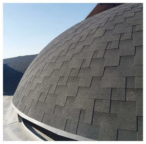 Detailný záber na kupolovú strechu pokrytú KATEPAL Oy Asfaltový strešný šindeľ Katepal Rocky in grafitová šedá, usporiadané v prekrývajúcom sa vzore na pozadí jasnej modrej oblohy.