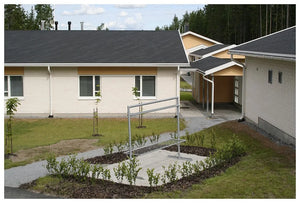 Malé vydláždené nádvorie s kovovou lavičkou a mladými stromčekmi je ohraničené nízkymi, svetlými budovami zastrešenými KATEPAL Oy Asfaltový strešný šindeľ Katepal Skalky v grafitovej šedi a bielo orámovanými oknami, v blízkosti lesa pod čiastočne zamračenou oblohou.