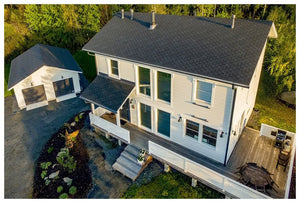 Letecký pohľad na moderný dvojpodlažný dom s KATEPAL Oy Asfaltový strešný šindeľ Katepal Rocky - grafitová šedá strecha, veľké okná, drevená terasa, samostatná garáž, obklopený zeleňou a štrkovou príjazdovou cestou.