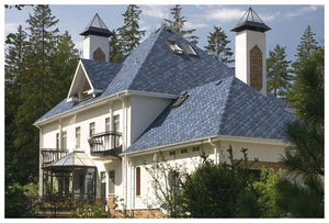 Priestranný dvojpodlažný dom so strmými strechami pokrytými Asfaltovým strešným šindeľom KATEPAL Oy Katepal Rocky - námornícka modrá, vysoké komíny, množstvo okien a balkónov, obklopený stromami pod čiastočne zamračenou oblohou.