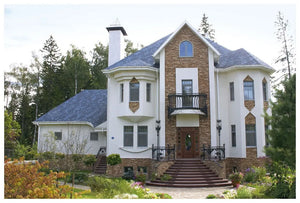 Priestranný dvojpodlažný dom s bielymi stenami, hnedými tehlovými akcentmi, oblúkovými oknami, balkónom, stĺpmi a upravenou záhradou. Na streche sa nachádza elegantný Asfaltový strešný šindeľ Katepal Rocky - námornícka modrá od spoločnosti KATEPAL Oy.