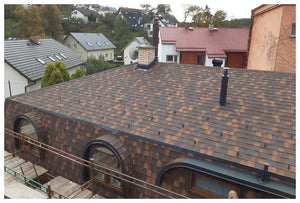 Pohľad na strechu budovy s KATEPAL Oy Asfaltový strešný šindeľ Katepal Mansion - medený, viaceré komíny a vetracie otvory, oblúkové okná; v pozadí sú viditeľné okolité obytné domy a zeleň.