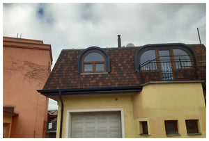 Žltý dom so strechou Asfaltový strešný šindeľ Katepal Mansion - medený od spoločnosti KATEPAL Oy má dve oblúkové okná (jedno s balkónom), viditeľné garážové dvere pod ním a oranžovú budovu vľavo pod čiastočne zamračenou oblohou.