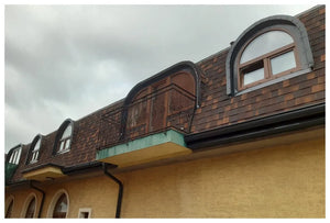 Budova s medeným Asfaltovým strešným šindľom Katepal Mansion od spoločnosti KATEPAL Oy a oblúkovými oknami, s malým balkónom s ozdobným železným zábradlím pod väčším oblúkovým oknom, pod zamračenou oblohou.