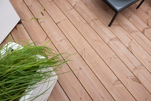 Detailný pohľad na drevenú palubu so svetlohnedými doskami. V ľavom dolnom rohu je kvetináč s vysokou zelenou trávou a v pravom hornom rohu je vidieť časť stoličky alebo stola s čiernymi nohami.